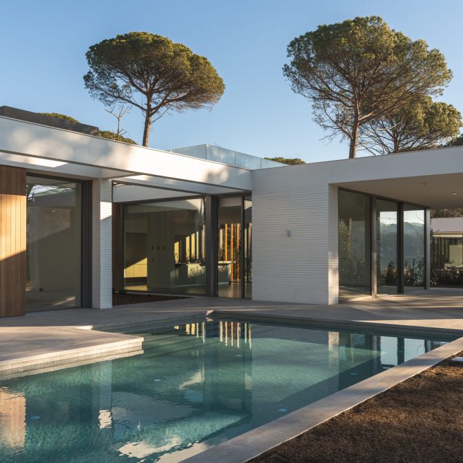 Outdoor view of a white and wooden luxury house with swimming pools and pine trees outside.