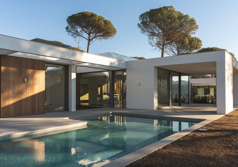Outdoor view of a white and wooden luxury house with swimming pools and pine trees outside.