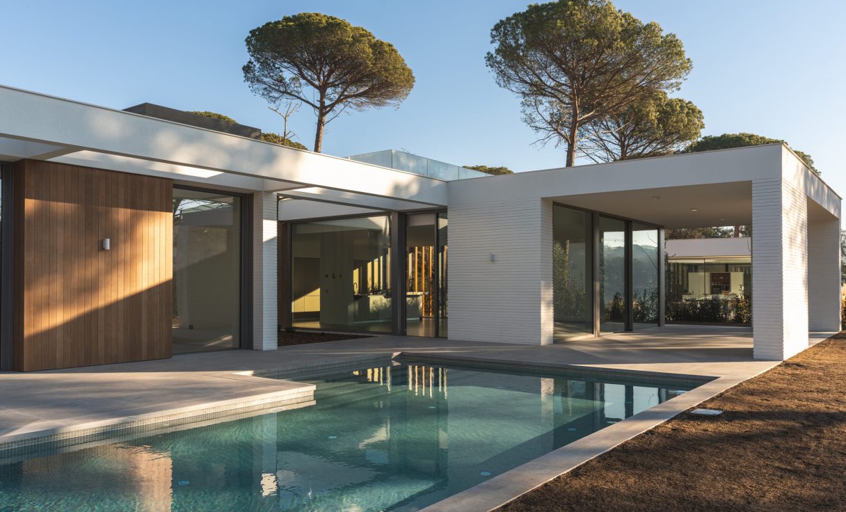 Outdoor view of a white and wooden luxury house with swimming pools and pine trees outside.